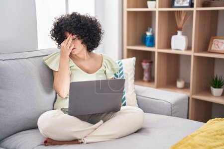 Foto de Young brunette woman with curly hair using laptop sitting on the sofa at home tired rubbing nose and eyes feeling fatigue and headache. stress and frustration concept. - Imagen libre de derechos