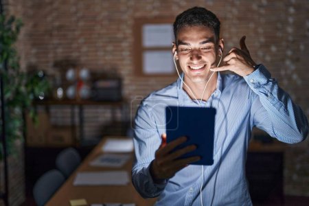Téléchargez les photos : Un bel homme hispanique travaillant au bureau la nuit souriant faisant un geste téléphonique avec les mains et les doigts comme parler au téléphone. communiquer les concepts. - en image libre de droit