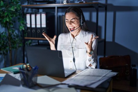 Photo for Young brunette woman wearing call center agent headset working late at night celebrating mad and crazy for success with arms raised and closed eyes screaming excited. winner concept - Royalty Free Image