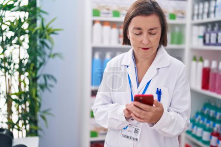Téléchargez les photos : Middle age woman pharmacist using smartphone working at pharmacy - en image libre de droit