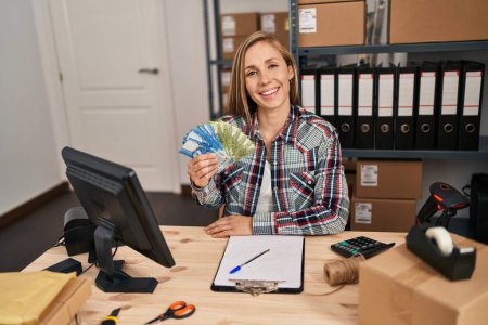 Foto de Young blonde woman working at small business ecommerce holding chilean pesos banknotes looking positive and happy standing and smiling with a confident smile showing teeth - Imagen libre de derechos