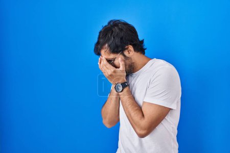 Foto de Hombre latino guapo de pie sobre fondo azul con expresión triste cubriendo la cara con las manos mientras llora. concepto de depresión. - Imagen libre de derechos