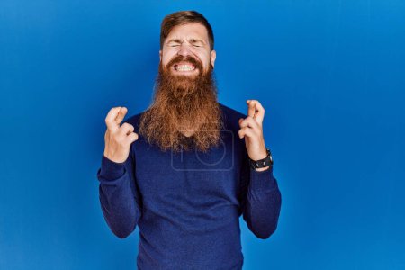 Téléchargez les photos : Homme rousse à longue barbe portant un pull bleu décontracté sur fond bleu geste doigt croisé souriant avec espoir et les yeux fermés. chance et concept superstitieux. - en image libre de droit
