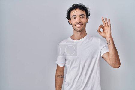 Foto de Hombre hispano de pie sobre fondo aislado sonriendo positivo haciendo signo de ok con la mano y los dedos. expresión exitosa. - Imagen libre de derechos