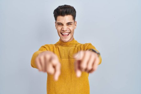 Foto de Young hispanic man standing over blue background pointing to you and the camera with fingers, smiling positive and cheerful - Imagen libre de derechos
