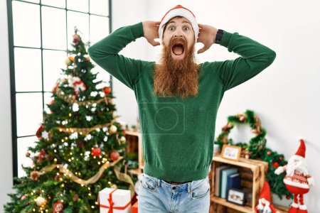 Téléchargez les photos : Homme rousse à longue barbe portant un chapeau de Noël par arbre de Noël fou et effrayé avec les mains sur la tête, effrayé et surpris de choc avec la bouche ouverte - en image libre de droit