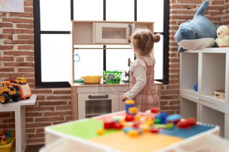 Foto de Adorable chica caucásica jugando con el juego de cocina de pie en la vista trasera en el jardín de infantes - Imagen libre de derechos