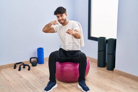 Foto de Hombre hispano con barba sentado en bolas de pilate en la sala de yoga señalando con los dedos a la cámara con cara alegre y divertida. buena energía y vibraciones. - Imagen libre de derechos