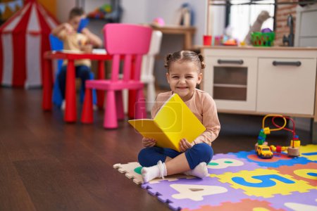 Photo for Adorable hispanic girl student sitting on floor reading book at kindergarten - Royalty Free Image