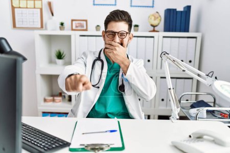 Téléchargez les photos : Young man with beard wearing doctor uniform and stethoscope at the clinic laughing at you, pointing finger to the camera with hand over mouth, shame expression - en image libre de droit
