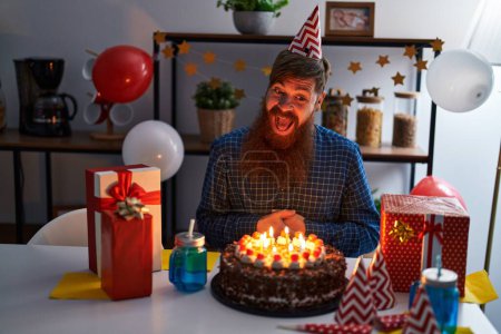 Foto de Young redhead man celebrating birthday sitting on table at home - Imagen libre de derechos