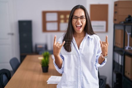 Téléchargez les photos : Jeune femme hispanique au bureau criant avec une expression folle faire symbole rock avec les mains en l'air. star de la musique. concept de musique lourde. - en image libre de droit