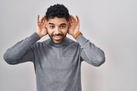 Photo for Hispanic man with beard standing over white background trying to hear both hands on ear gesture, curious for gossip. hearing problem, deaf - Royalty Free Image