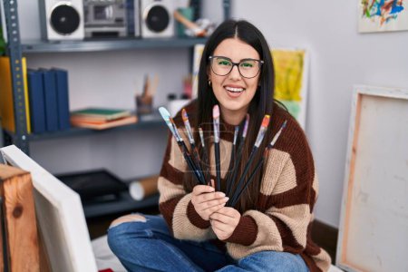 Photo for Young hispanic woman smiling confident holding paintbrushes at art studio - Royalty Free Image