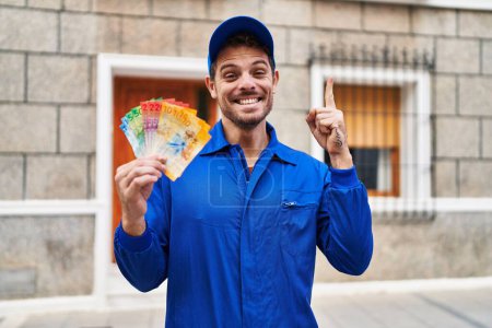 Téléchargez les photos : Jeune homme hispanique portant un uniforme de bricoleur tenant des francs suisses souriant avec une idée ou une question pointant du doigt avec un visage heureux, numéro un - en image libre de droit