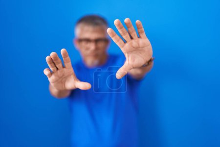 Foto de Hombre hispano con el pelo gris de pie sobre fondo azul haciendo marco usando las manos palmas y los dedos, perspectiva de la cámara - Imagen libre de derechos