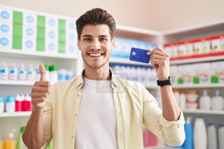 Foto de Young hispanic man at pharmacy drugstore holding credit card smiling happy and positive, thumb up doing excellent and approval sign - Imagen libre de derechos