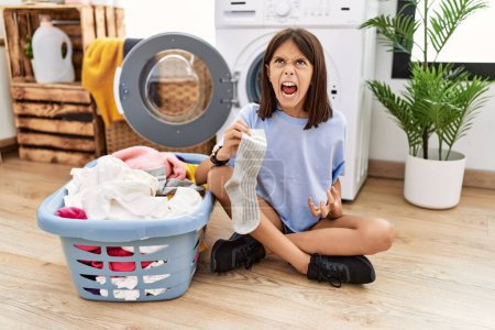 Foto de Young hispanic girl doing laundry holding socks angry and mad screaming frustrated and furious, shouting with anger. rage and aggressive concept. - Imagen libre de derechos