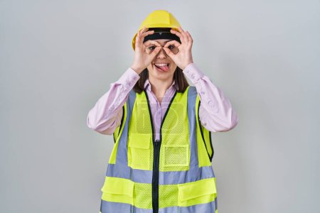 Photo for Hispanic girl wearing builder uniform and hardhat doing ok gesture like binoculars sticking tongue out, eyes looking through fingers. crazy expression. - Royalty Free Image