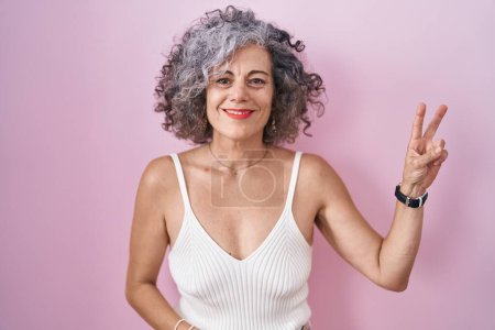 Téléchargez les photos : Middle age woman with grey hair standing over pink background smiling with happy face winking at the camera doing victory sign with fingers. number two. - en image libre de droit