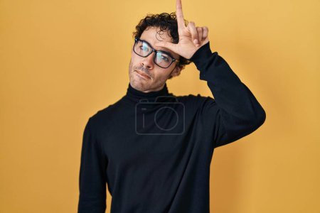 Photo for Hispanic man standing over yellow background making fun of people with fingers on forehead doing loser gesture mocking and insulting. - Royalty Free Image