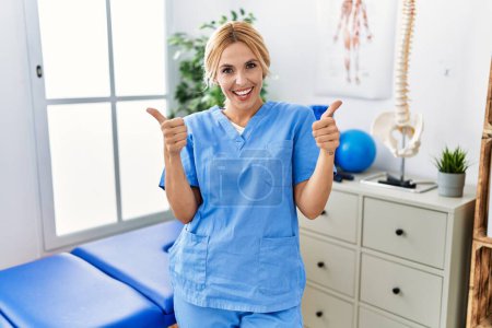 Photo for Beautiful blonde physiotherapist woman working at pain recovery clinic success sign doing positive gesture with hand, thumbs up smiling and happy. cheerful expression and winner gesture. - Royalty Free Image
