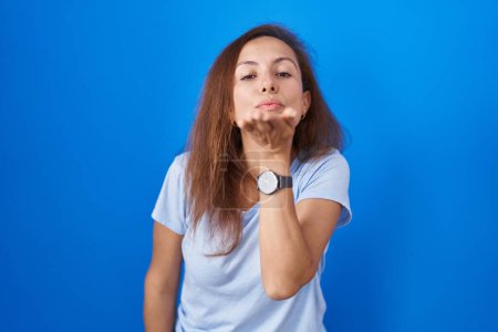 Foto de Brunette woman standing over blue background looking at the camera blowing a kiss with hand on air being lovely and sexy. love expression. - Imagen libre de derechos