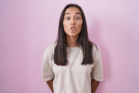 Foto de Mujer hispana joven de pie sobre fondo rosa haciendo cara de pez con labios, gesto loco y cómico. expresión divertida. - Imagen libre de derechos