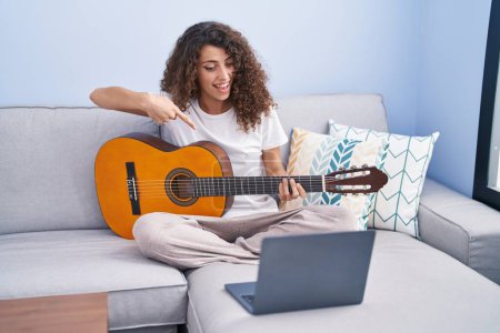 Téléchargez les photos : Jeune belle femme hispanique ayant cours de guitare classique en ligne assis sur le canapé à la maison - en image libre de droit