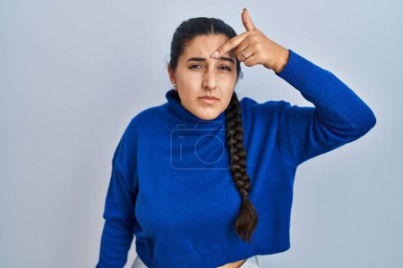 Foto de Young hispanic woman standing over isolated background pointing unhappy to pimple on forehead, ugly infection of blackhead. acne and skin problem - Imagen libre de derechos