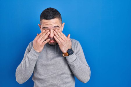 Foto de Hispanic man standing over blue background rubbing eyes for fatigue and headache, sleepy and tired expression. vision problem - Imagen libre de derechos