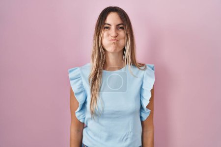 Foto de Young hispanic woman standing over pink background puffing cheeks with funny face. mouth inflated with air, crazy expression. - Imagen libre de derechos