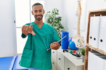Foto de Hombre fisioterapeuta afroamericano que trabaja en la clínica de recuperación del dolor señalándote a ti y a la cámara con los dedos, sonriendo positiva y alegre - Imagen libre de derechos