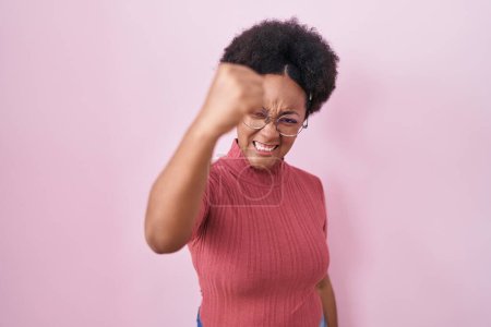 Photo for Beautiful african woman with curly hair standing over pink background angry and mad raising fist frustrated and furious while shouting with anger. rage and aggressive concept. - Royalty Free Image