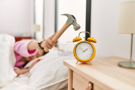 Foto de Chica apagando despertador usando martillo acostado en la cama en el dormitorio. - Imagen libre de derechos
