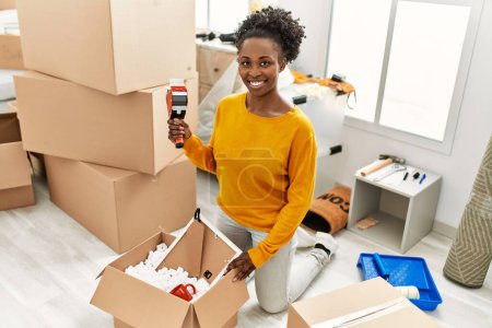 Photo for African american woman smiling confident packing cardboard box at new home - Royalty Free Image
