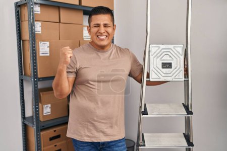Téléchargez les photos : Jeune homme hispanique travaillant dans le commerce électronique de petite entreprise tenant échelles criant fier, célébrant la victoire et le succès très excité avec bras levé - en image libre de droit