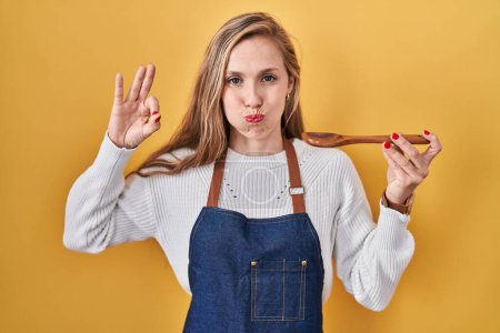 Foto de Young blonde woman wearing apron tasting food holding wooden spoon puffing cheeks with funny face. mouth inflated with air, catching air. - Imagen libre de derechos
