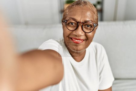 Téléchargez les photos : Femme afro-américaine senior faire selfie par la caméra assis sur le canapé à la maison - en image libre de droit