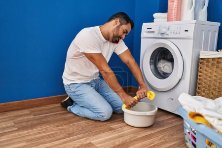 Photo for Young hispanic man talking on the smartphone wringing out rag at laundry room - Royalty Free Image