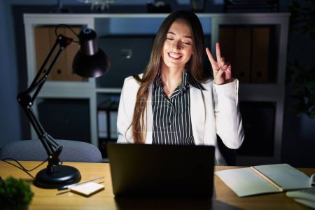 Téléchargez les photos : Jeune femme brune travaillant au bureau la nuit avec un ordinateur portable souriant regardant la caméra montrant des doigts faisant signe de victoire. numéro deux. - en image libre de droit