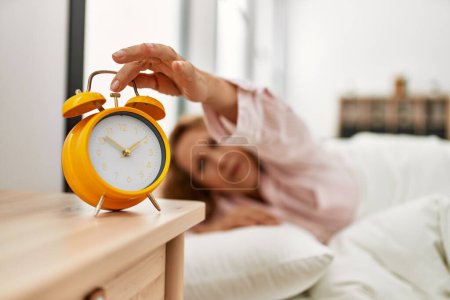 Foto de Middle age caucasian woman turning off alarm clock lying on the bed at bedroom. - Imagen libre de derechos