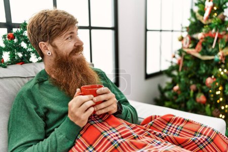 Photo for Young redhead man drinking coffee sitting by christmas tree at home - Royalty Free Image