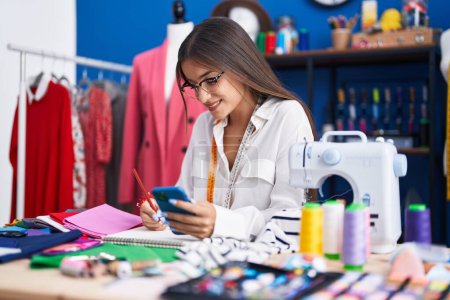 Téléchargez les photos : Jeune belle femme hispanique tailleur en utilisant le dessin smartphone sur ordinateur portable à l'usine de vêtements - en image libre de droit