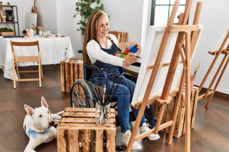 Téléchargez les photos : Jeune femme caucasienne souriante confiante assise sur un fauteuil roulant dessin avec chien au studio d'art - en image libre de droit