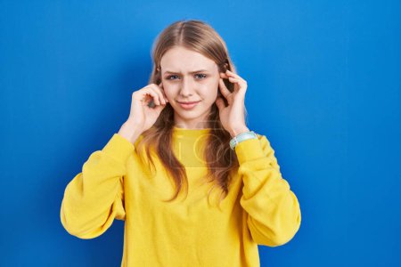 Téléchargez les photos : Jeune femme caucasienne debout sur fond bleu couvrant les oreilles avec des doigts avec une expression agacée pour le bruit de la musique forte. concept sourd. - en image libre de droit