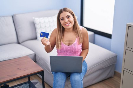 Foto de Joven mujer hispana hermosa usando portátil y tarjeta de crédito sentado en el sofá en casa - Imagen libre de derechos