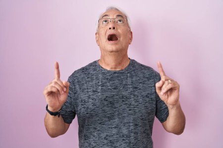 Foto de Hombre de mediana edad con el pelo gris de pie sobre fondo rosa asombrado y sorprendido mirando hacia arriba y señalando con los dedos y los brazos levantados. - Imagen libre de derechos
