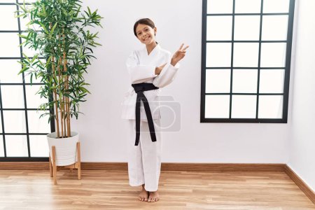 Photo for Young hispanic girl wearing karate kimono and black belt smiling with happy face winking at the camera doing victory sign. number two. - Royalty Free Image