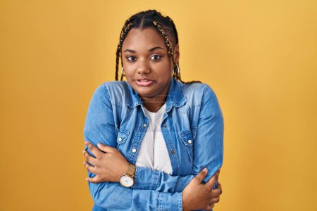 Foto de African american woman with braids standing over yellow background shaking and freezing for winter cold with sad and shock expression on face - Imagen libre de derechos